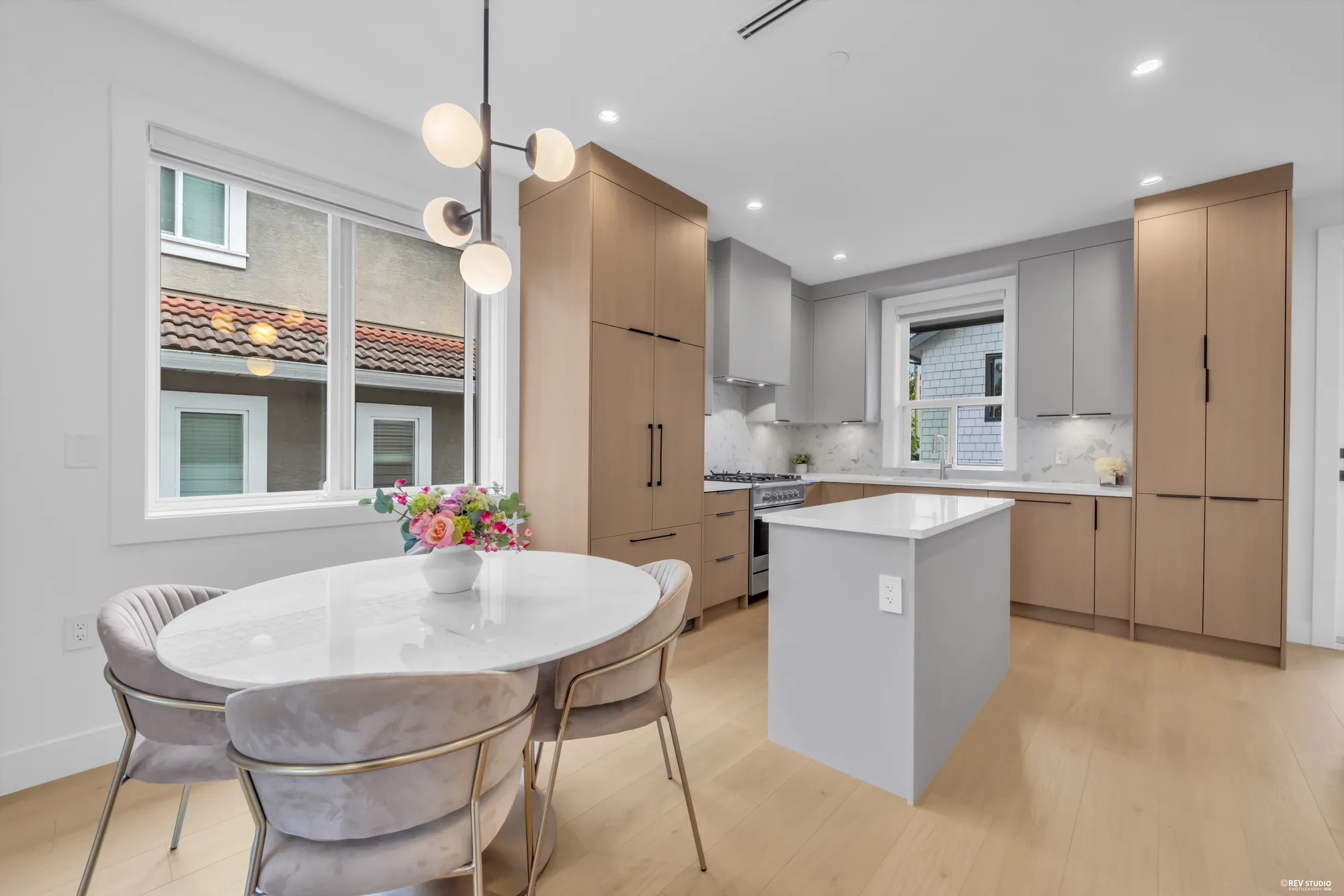 Round dining table with upholstered chairs beside a window, kitchen island behind