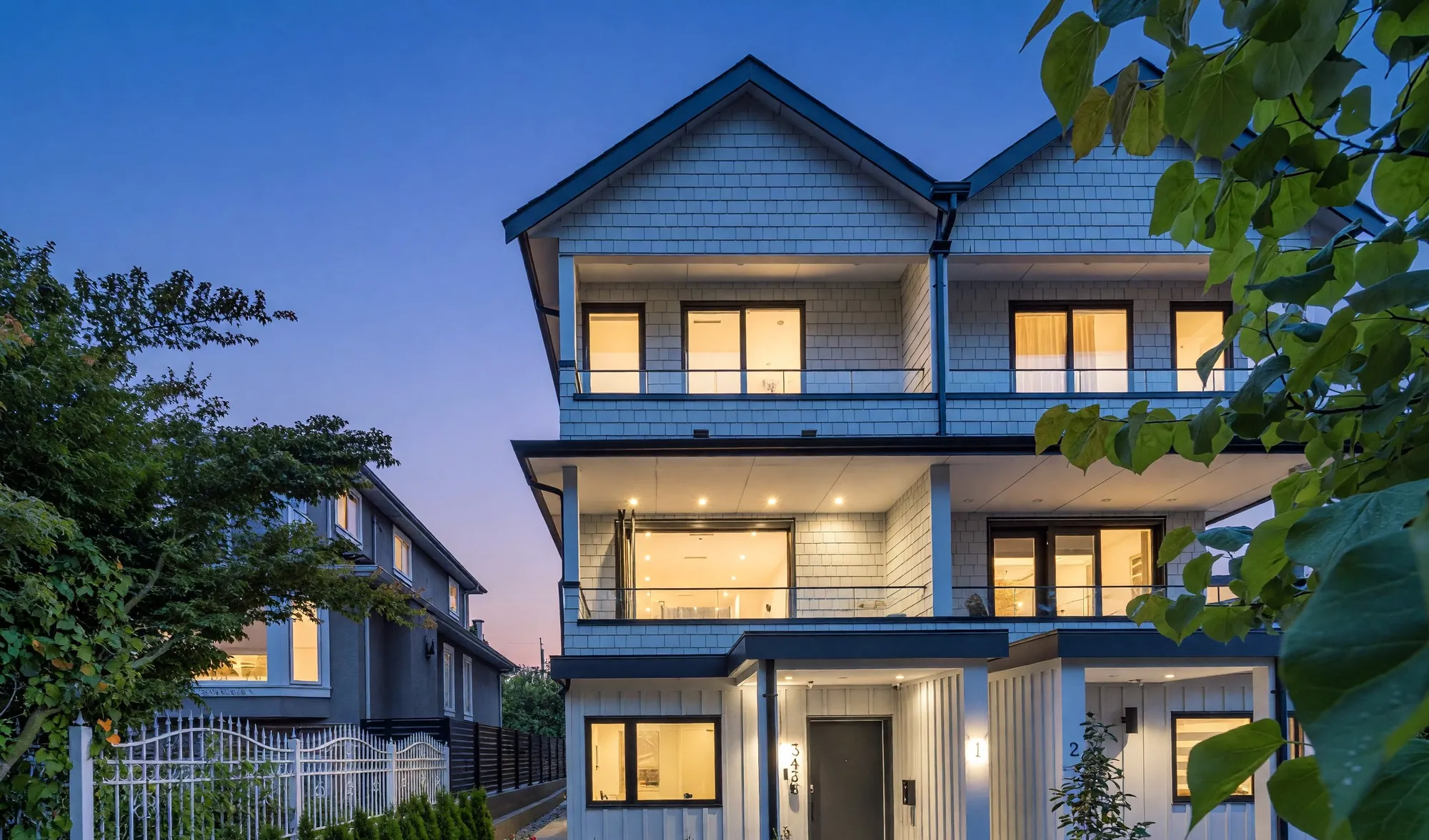 1-3436 Triumph Street at dusk: a new three-storey house with lit windows, glass balcony rails and a low black fence along the sidewalk
